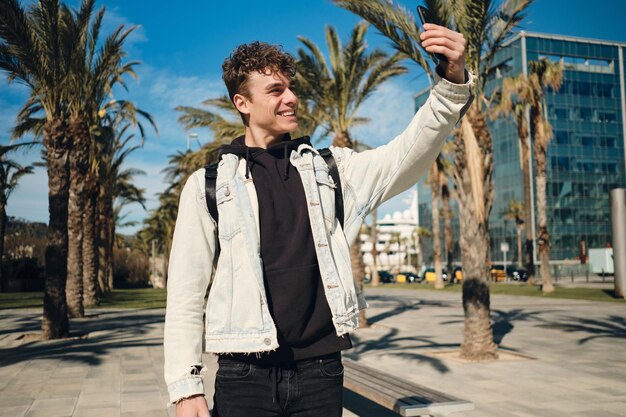 Handsome smiling guy happily taking selfie on smartphone in city park. Young stylish tourist man walking outdoor