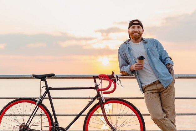 Handsome smiling happy hipster style bearded man wearing denim shirt and cap with bicycle in morning sunrise by the sea drinking coffee, healthy active lifestyle traveler