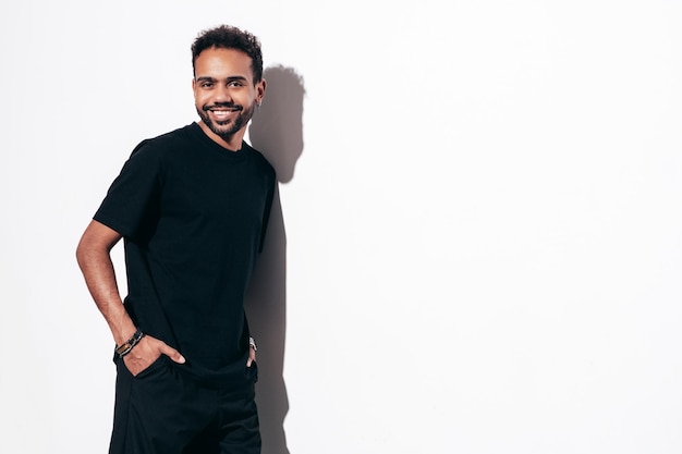 Handsome smiling hipster model Sexy unshaven man dressed in black summer tshirt and jeans clothes Fashion male posing near white wall in studio at sunny day Isolated