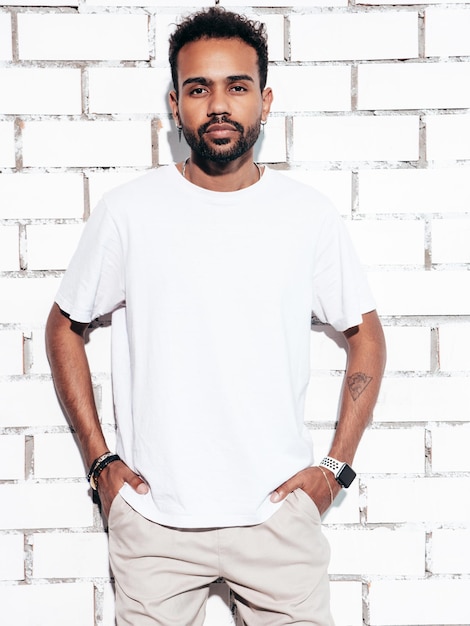 Handsome smiling hipster model Sexy unshaven man dressed in white summer tshirt and jeans clothes Fashion male posing near white brick wall in studio at sunny day Isolated