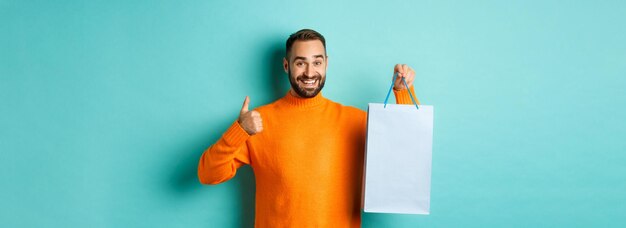 Handsome smiling man showing thumbsup and shopping bag recommending store standing over blue backgro