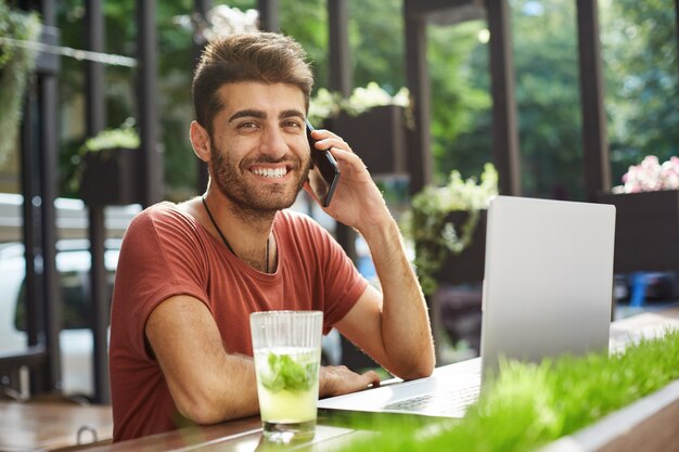 Handsome smiling man talking on mobile phone, calling to seller he found online while using laptop, shopping, searching appartment in internet