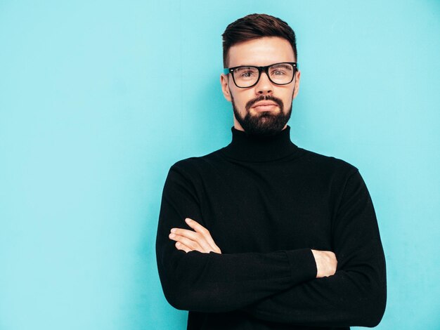 Handsome smiling model Sexy stylish man dressed in black turtleneck sweater and jeans Fashion hipster male posing near blue wall in studio Isolated In spectacles or eyeglasses Crossed arms