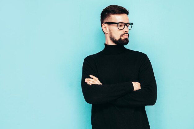 Handsome smiling model Sexy stylish man dressed in black turtleneck sweater and jeans Fashion hipster male posing near blue wall in studio Isolated In spectacles or eyeglasses Crossed arms