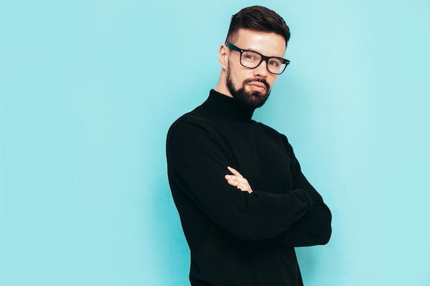 Handsome smiling model Sexy stylish man dressed in black turtleneck sweater and jeans Fashion hipster male posing near blue wall in studio Isolated In spectacles or eyeglasses Crossed arms