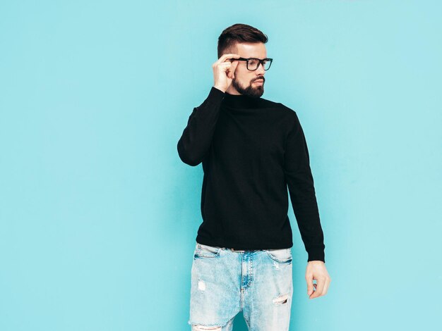 Handsome smiling model Sexy stylish man dressed in black turtleneck sweater and jeans Fashion hipster male posing near blue wall in studio Isolated