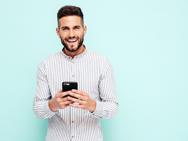 Handsome smiling modelSexy stylish man dressed in shirt and jeans Fashion hipster male posing near blue wall in studio Holding smartphone Looking at cellphone screen Using apps