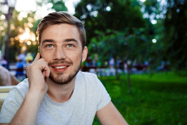 Handsome smiling young man sitting at the table in open-air cafe talkin on phone looking up.