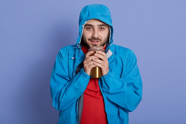 Free Photo handsome smiling young man with thermo cup in hands, keeping warm, drinking hot beverage, looks at camera, wearing blue jacket