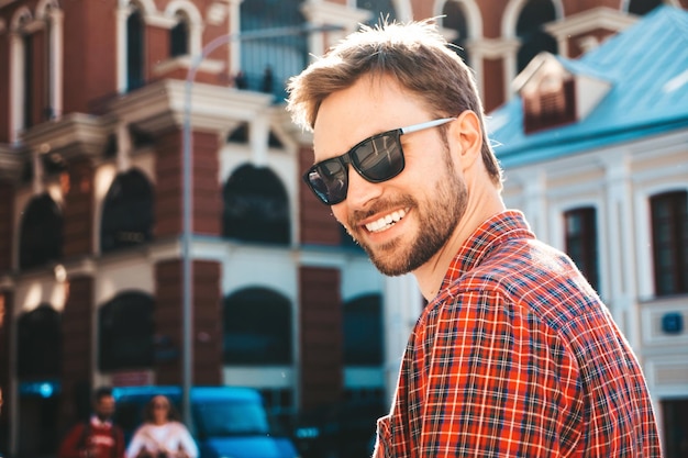 Free photo handsome stylish hipster lambersexual model.modern man dressed in red checkered shirt. fashion male posing near skyscraper on the street background in sunglasses. outdoors at sunset