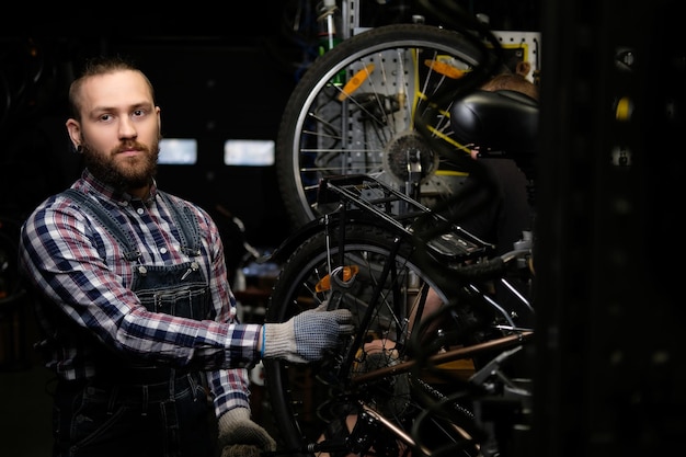 A handsome stylish male wearing a flannel shirt and jeans coverall, working with a bicycle wheel in a repair shop. A worker using a wrench mounts the wheel on a bike in a workshop.