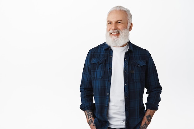 Handsome stylish old hipster man senior with white beard and tattoos looking aside at advertisement with pleased face smiling happy reading promotional text standing over studio background