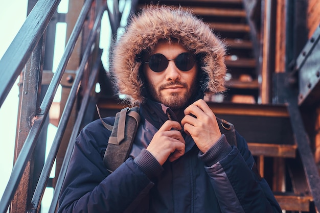 Free Photo a handsome stylish young man wearing a winter coat and sunglasses sitting on stairs outside.