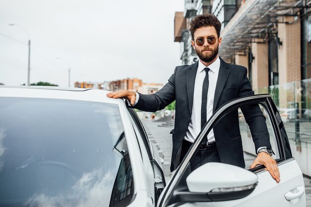 A handsome successful businessman in full suit opens his car on the streets of the city near the modern office center