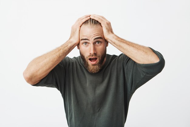 Handsome swedish man with trendy hairstyle and beard holding hands on head shocked