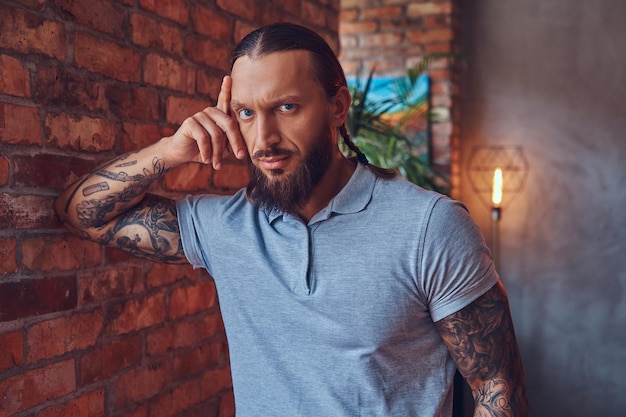 Free photo handsome tattoed male with a stylish haircut and beard, in a gray t-shirt, standing leaning against a brick wall in a room with a loft interior.