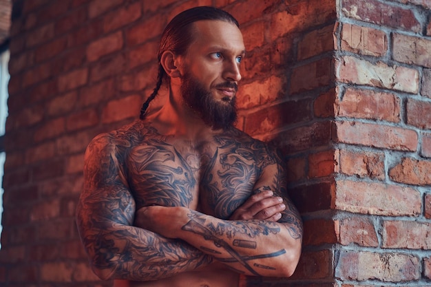 Free photo handsome tattoed shirtless male with a stylish haircut and beard, standing with crossed arms, leaning against a brick wall in a room with a loft interior.