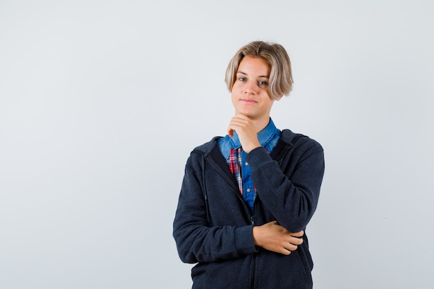 Handsome teen boy in shirt, hoodie propping chin on hand and looking cheerful , front view.