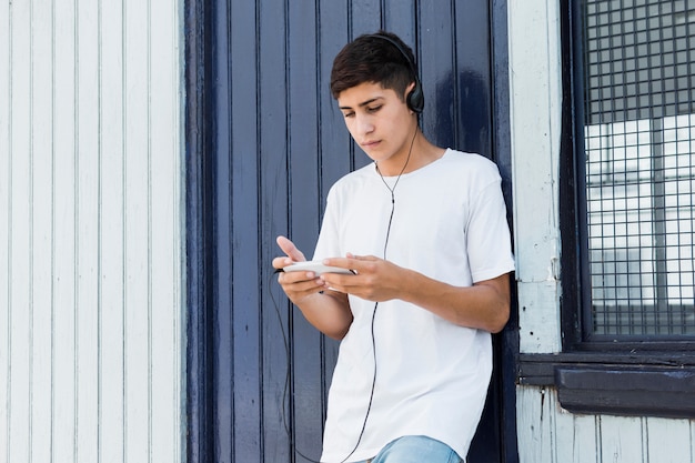 Handsome teenage boy leaning on metal wall using smartphone and listening music