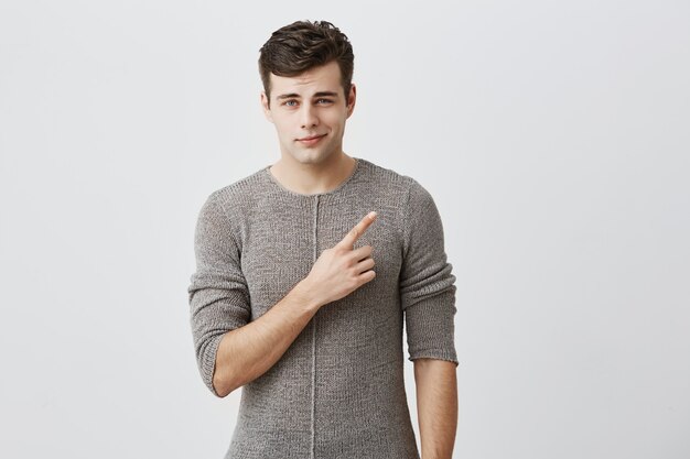 Handsome trendy man wearing casual clothes, smiling at corner of his mouth, dressed casually, pointing with forefinger aside at text or advertisment. Male model posing.