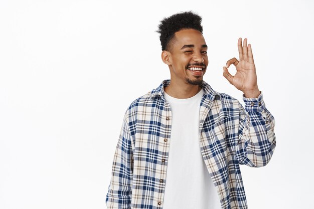 handsome young african american guy smiling satisfied, showing OK, okay zero sign and nod in approval, rate good thing, praise excellent work, recommend store, standing on white