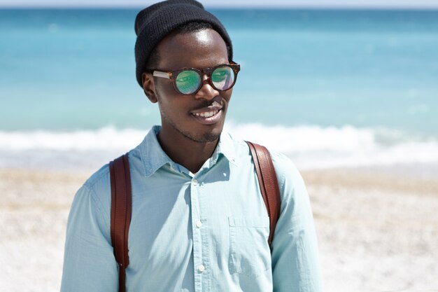 Handsome young African American hipster walking along coast, admiring good weather and marine view, standing with his back to vast azure ocean