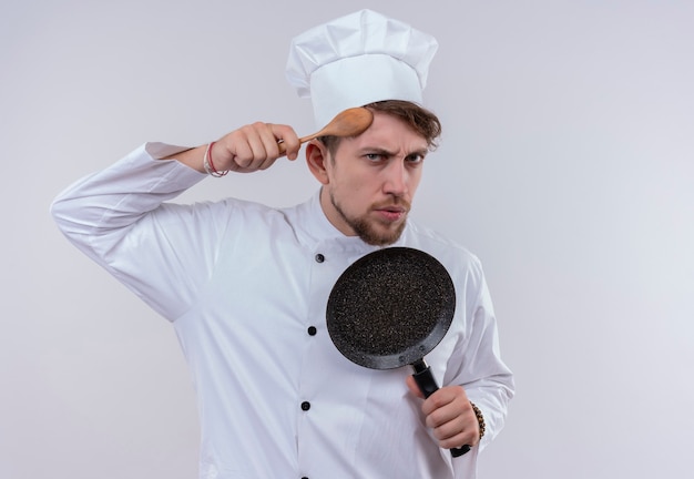 Free photo a handsome young bearded chef man wearing white cooker uniform and hat showing frying pan while holding wooden spoon on his head on a white wall