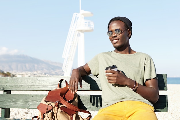 Handsome young black European male traveler dressed in trendy clothing having a few minutes rest on bench, drinking tea or coffee out of paper cup during long walk around resort town at daytime