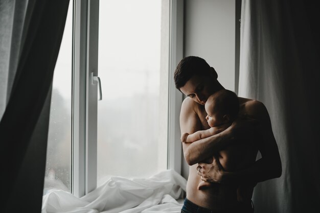 Handsome young dad holds newborn baby close to his heart standing before a window 