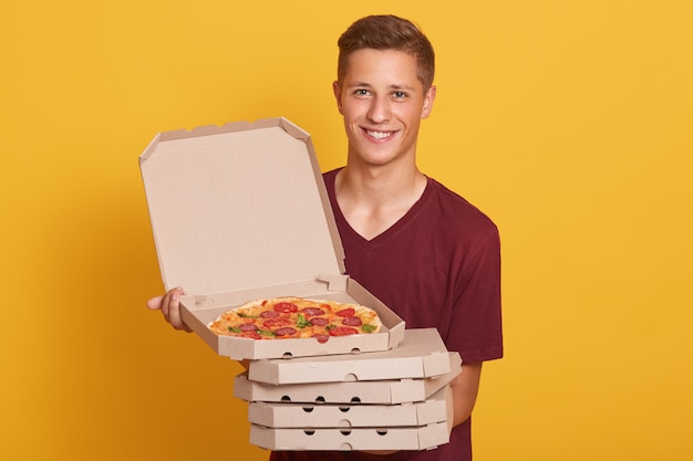 Free photo handsome young delivery worker holding stack of pizza boxes, dressed casual t shirt, looking at camera and smiling, showing open box with tasty pepperoni, posing isolated on yellow studio