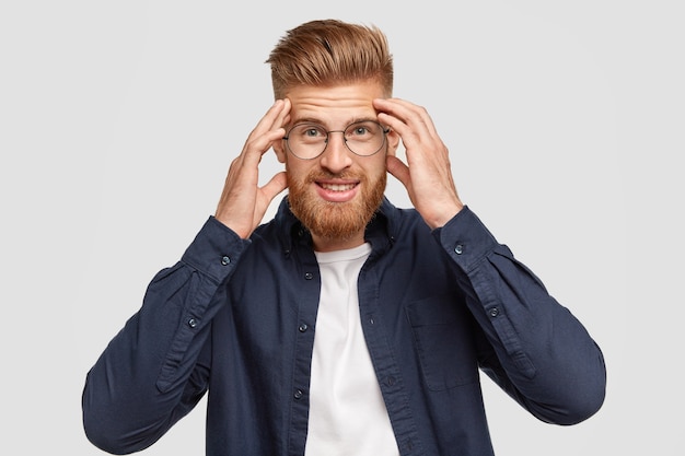 Handsome young ginger male keeps hands on temples, tries to concentrate on something, stands against white wall, has gentle smile, satisfied with results of work, finds solution in mind