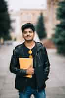 Free photo handsome young indian student man holding notebooks while standing on the street