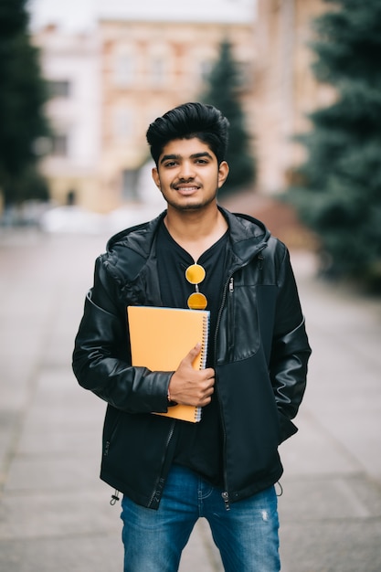 Free photo handsome young indian student man holding notebooks while standing on the street