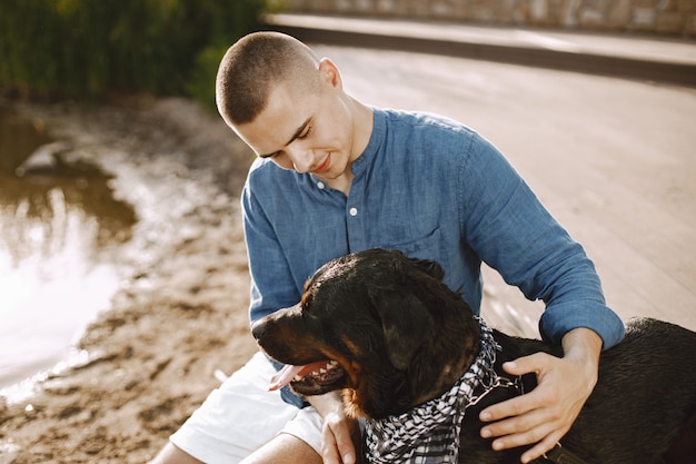 Free Photo handsome young male in casual outfit playing with cute dog while sitting near the lake. boy wearing blue shirt and white jeans shorts