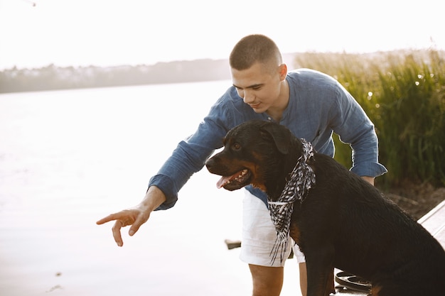 Free photo handsome young male in casual outfit playing with cute dog while standing near the lake. boy wearing blue shirt and white jeans shorts