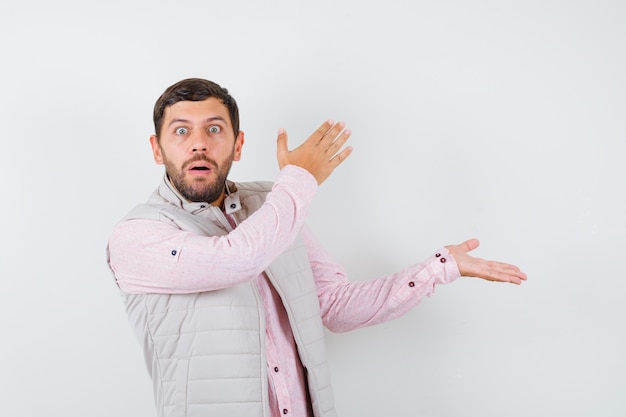 Handsome young male pretending to show something in shirt, vest and looking shocked , front view.