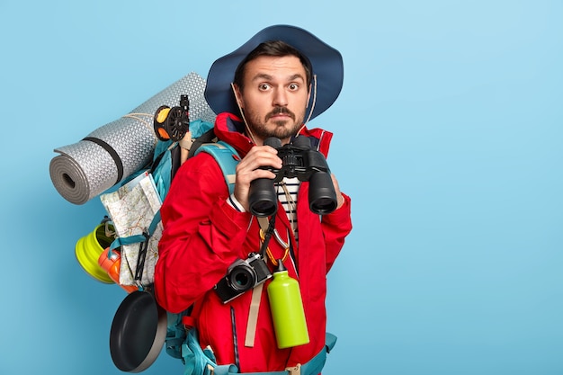 Free photo handsome young male tourist holds binoculars, walks in forest, wears casual clothes, carries necessary things for traveling, likes to spend holidays in active wear