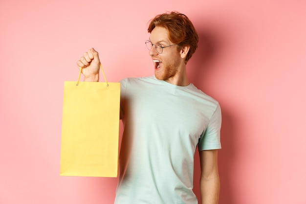 Handsome young man buying presents, holding shopping bag and looking amused, standing over pink background