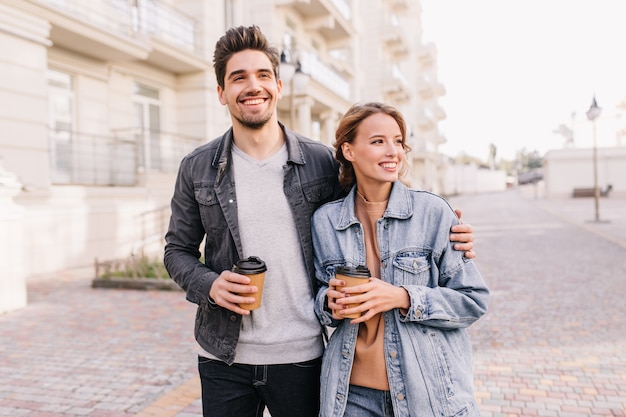 Handsome young man holding cup of coffee and embracing girlfriend. Smiling couple enjoying outdoor date.