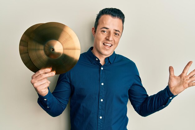 Handsome young man holding golden cymbal plates celebrating achievement with happy smile and winner expression with raised hand