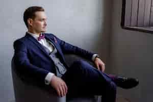 Free photo handsome young man is sitting in the armchair in the room, dressed in the fashionable suit