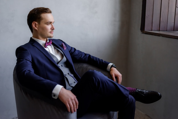 Free Photo handsome young man is sitting in the armchair in the room, dressed in the fashionable suit