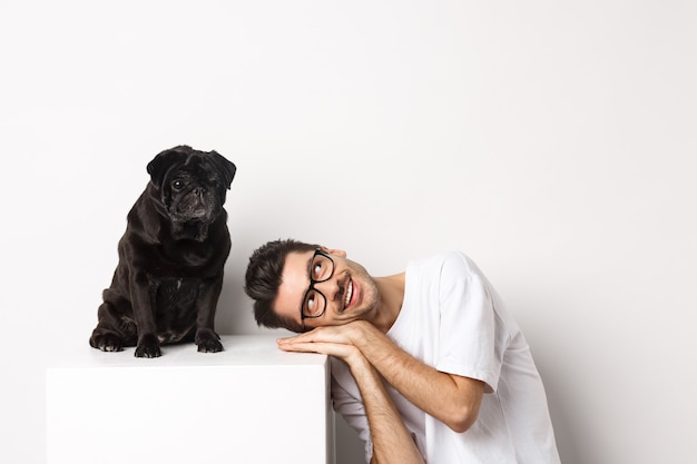 Free Photo handsome young man lay head near cute black pug, smiling and looking up at copy space, white background