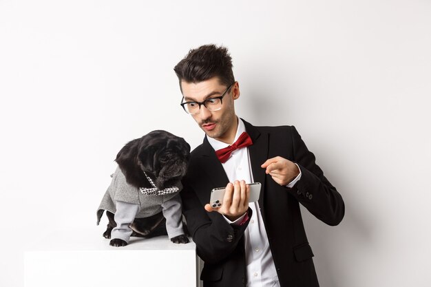 Handsome young man showing something on mobile phone to his dog. Owner shopping online with pet, standing in costumes over white background.