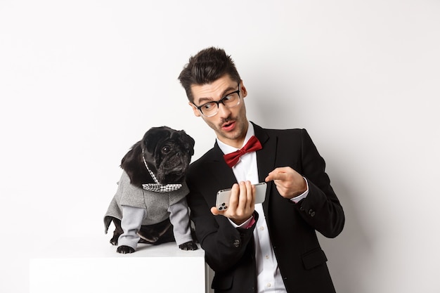 Free Photo handsome young man showing something on mobile phone to his dog. owner shopping online with pet, standing in costumes over white background