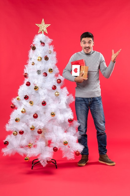 Handsome young man standing near the decorated white New Year tree and holding his gifts