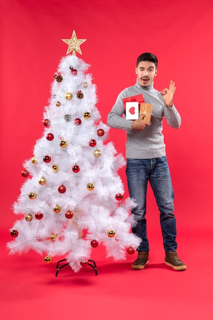 Handsome young man standing near the decorated white New Year tree and holding his gifts