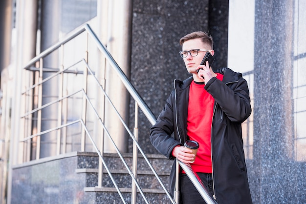 Handsome young man standing on skateboard on mobile phone holding takeaway coffee cup