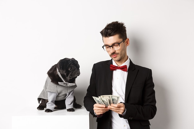 Free photo handsome young man in suit standing near black pug in costume and counting money, working on parties, posing over white background.