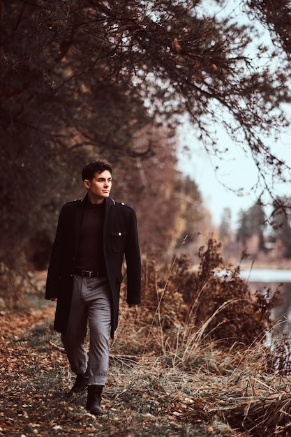 Free Photo a handsome young man wearing a black coat walking near a lake in the autumn forest.